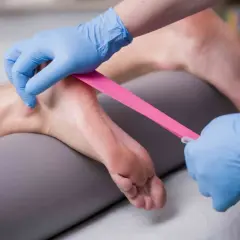Fizjoterapeuta w niebieskich rękawiczkach aplikuje różową taśmę kinezjotapingową na stopę pacjenta, wspierając rehabilitację.