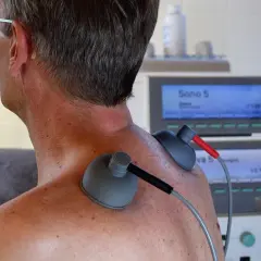 Pacjent z naciągniętymi więzadłami szyjnymi poddaje się terapii ultradźwiękami. Elektrody przymocowane do karku i ramienia.