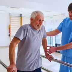 Starszy mężczyzna z kontuzją barku, pod opieką fizjoterapeuty, ćwiczy chodzenie przy poręczach. Długość przerwy zależy od postępów rehabilitacji.