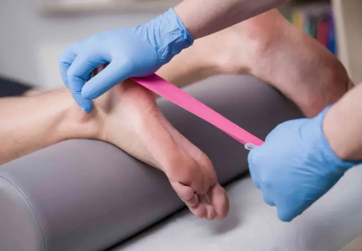 Fizjoterapeuta w niebieskich rękawiczkach aplikuje różową taśmę kinezjotapingową na stopę pacjenta, wspierając rehabilitację.