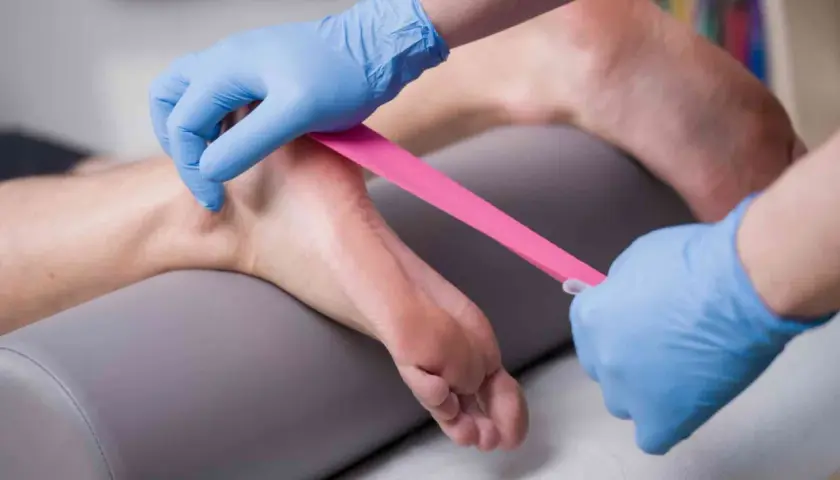 Fizjoterapeuta w niebieskich rękawiczkach aplikuje różową taśmę kinezjotapingową na stopę pacjenta, wspierając rehabilitację.