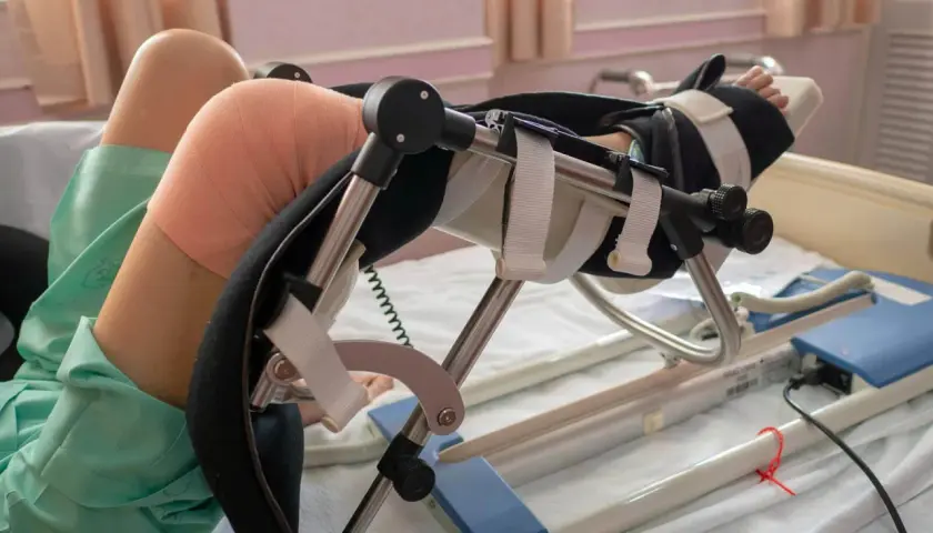 Pacjent w łóżku z nogą w aparacie do rehabilitacji po endoprotezie kolana.