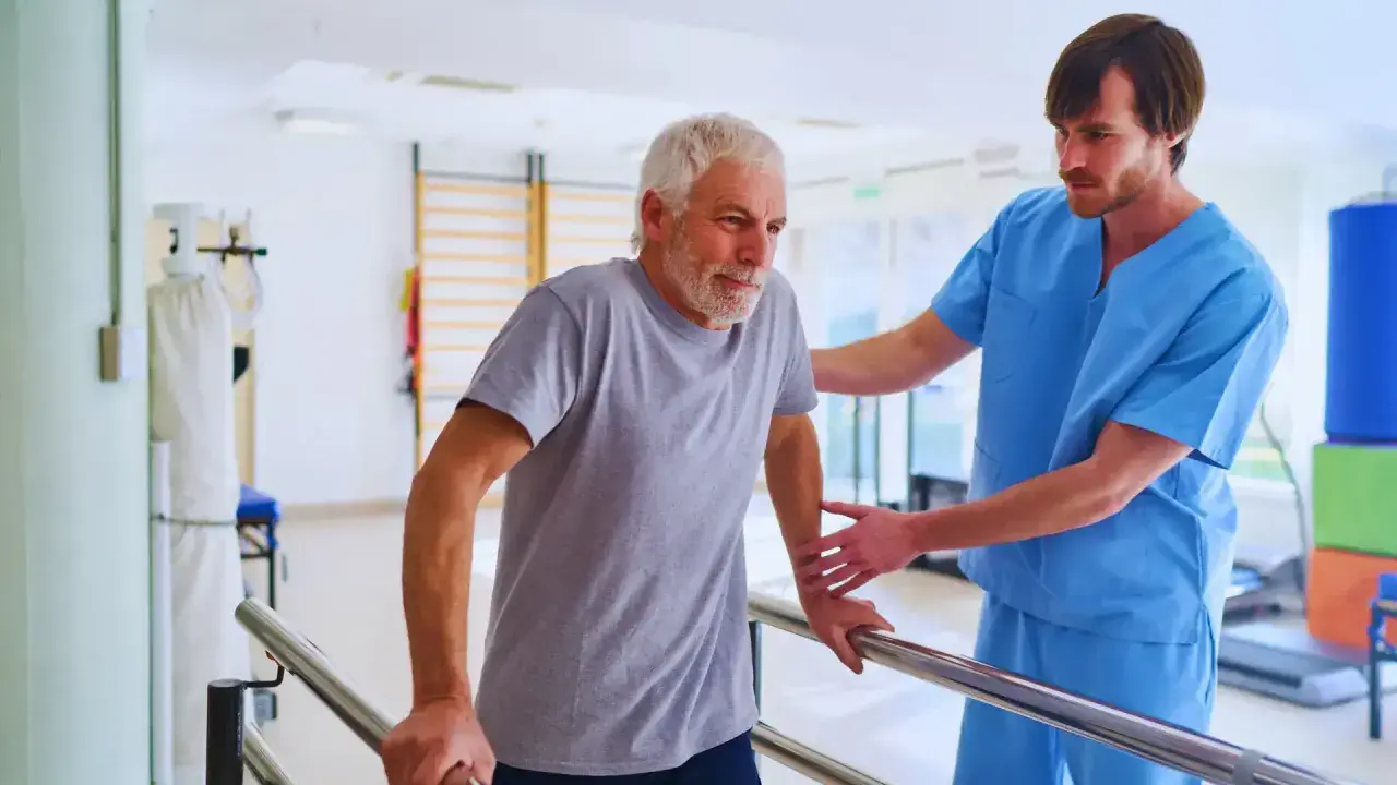 Starszy mężczyzna z kontuzją barku, pod opieką fizjoterapeuty, ćwiczy chodzenie przy poręczach. Długość przerwy zależy od postępów rehabilitacji.