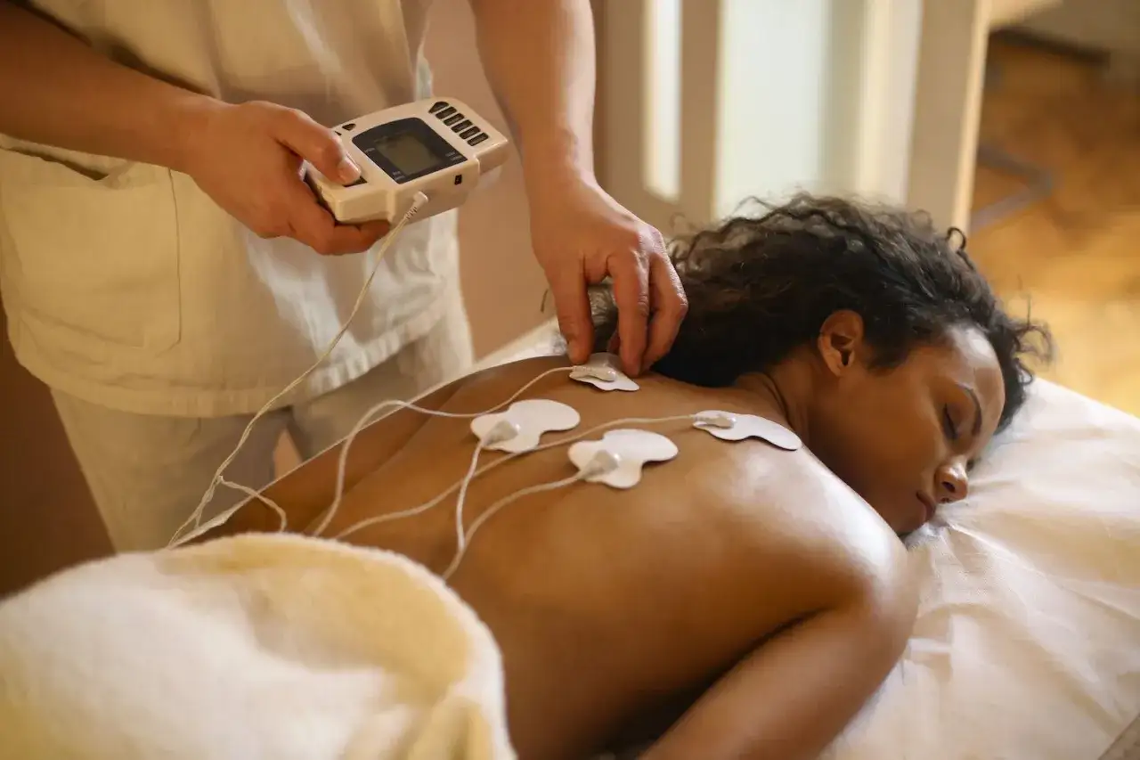 Kobieta leży na łóżku, a na jej plecach umieszczono elektrody. Terapeuta trzyma urządzenie do elektrostymulacji, oferując relaksujący zabieg w domu.